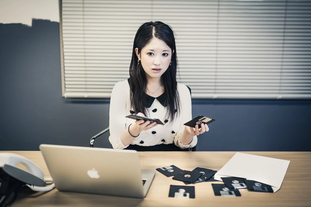  A young woman in a white blouse and black skirt sits at her desk, looking at a stack of 3.5-inch floppy disks with a laptop on the desk in front of her.